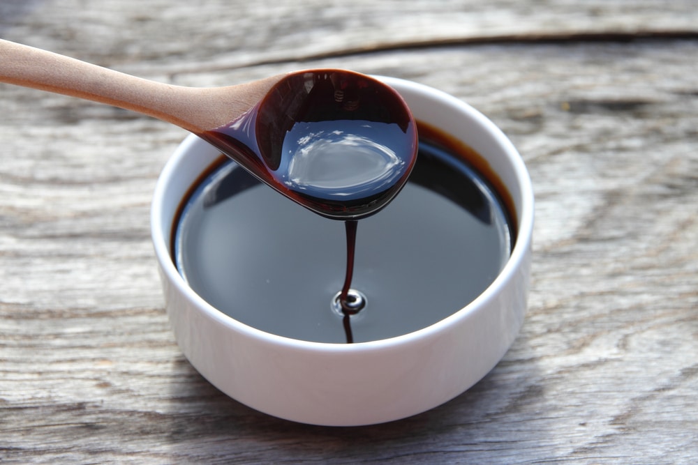 A white bowl filled with molasses on a light wood background. There is a wooden spoon with molasses in it, dripping into the bowl. 